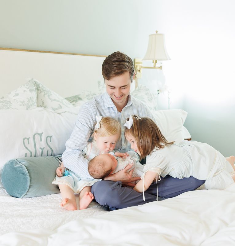 A smiling father sits on a white bed holding a newborn baby while two young siblings lean in to look.