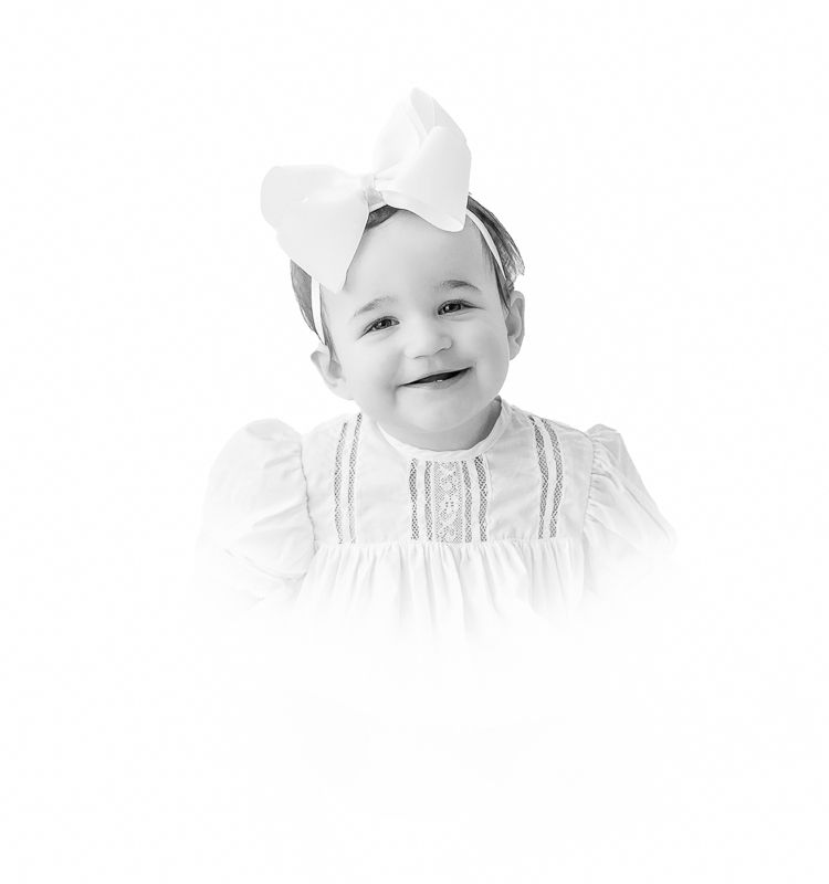 Black and white portrait of a smiling toddler girl wearing a white dress and large bow headband.