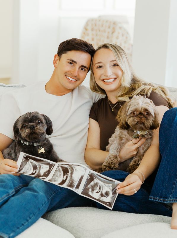 Smiling couple sitting on couch holding ultrasound photos with two small dogs, announcing pregnancy.