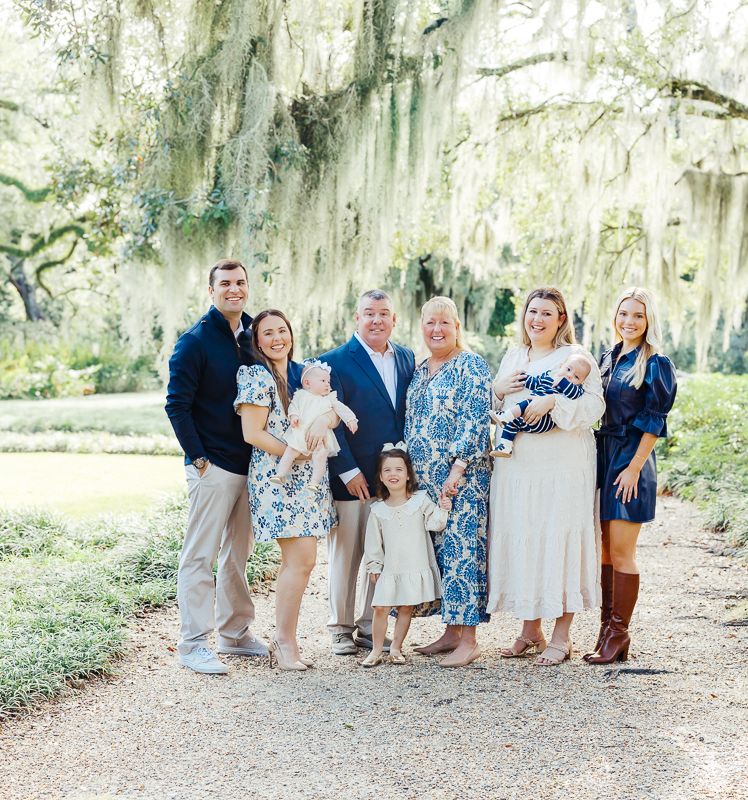 A multigenerational family poses together outdoors under moss-draped oak trees, dressed in elegant blue and white outfits.
