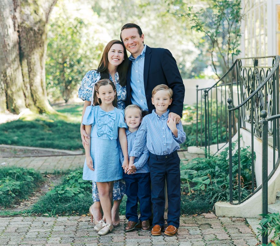 A happy family of five poses together on a brick path surrounded by lush greenery near iron stairs.