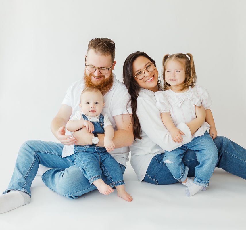 Happy young family with two children sitting together in white and denim outfits on white backdrop.