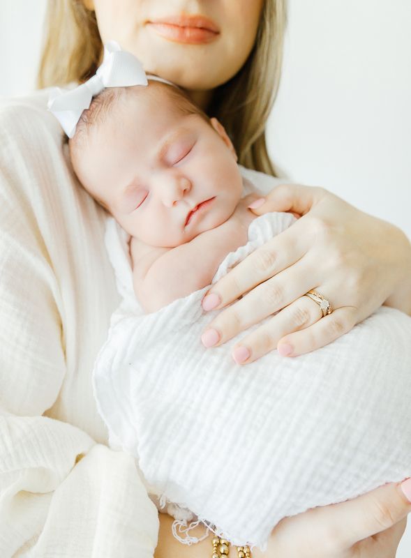 A mother gently holds her sleeping newborn baby wrapped in a white swaddle with a bow headband.