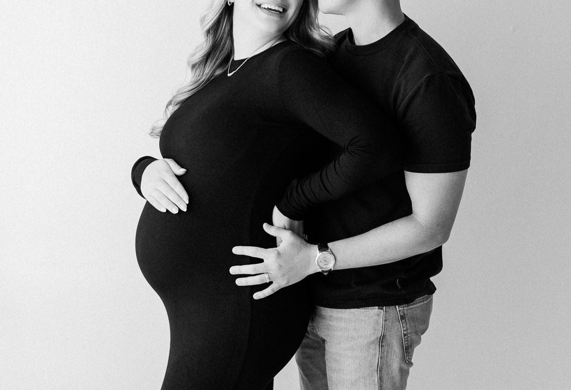 Black and white maternity photo of a couple, woman in black dress cradling baby bump, man embracing her from behind.
