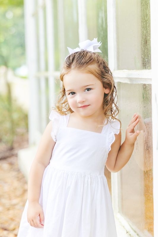 Young girl in white dress and bow poses by window in soft natural light.