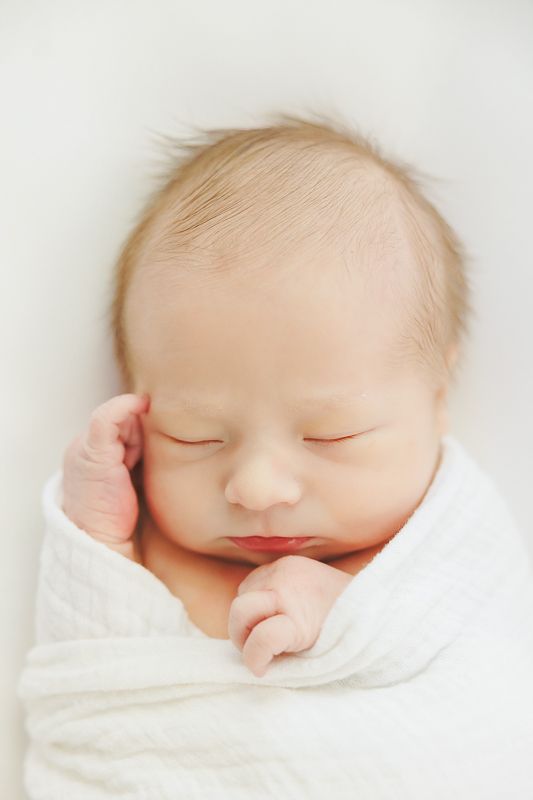 A peaceful newborn baby sleeping soundly, swaddled in a soft white blanket with tiny hand visible near face.
