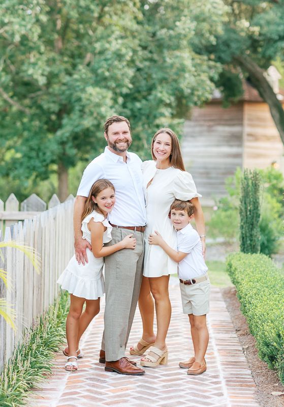 Happy family of four posing on a brick path in matching white outfits surrounded by lush greenery.