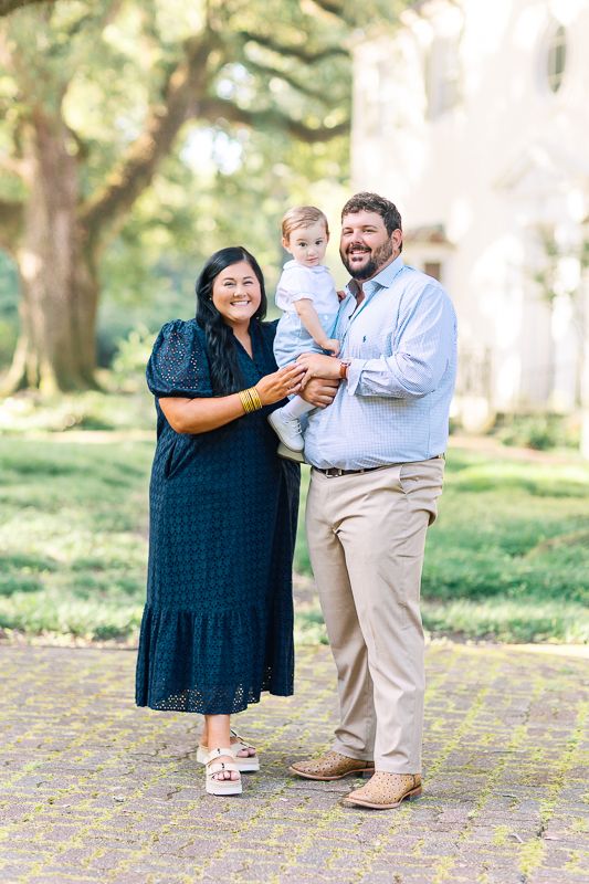 A smiling family of three poses outdoors, with a man holding a baby and a woman in a navy dress standing beside them.