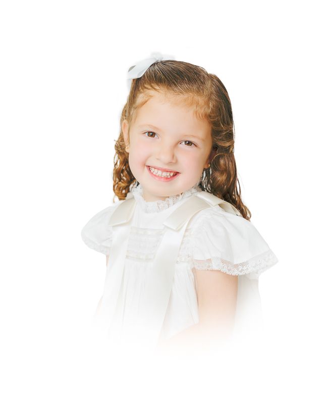 Smiling young girl with curly auburn hair wearing a white dress and bow, portrait photo on white background.