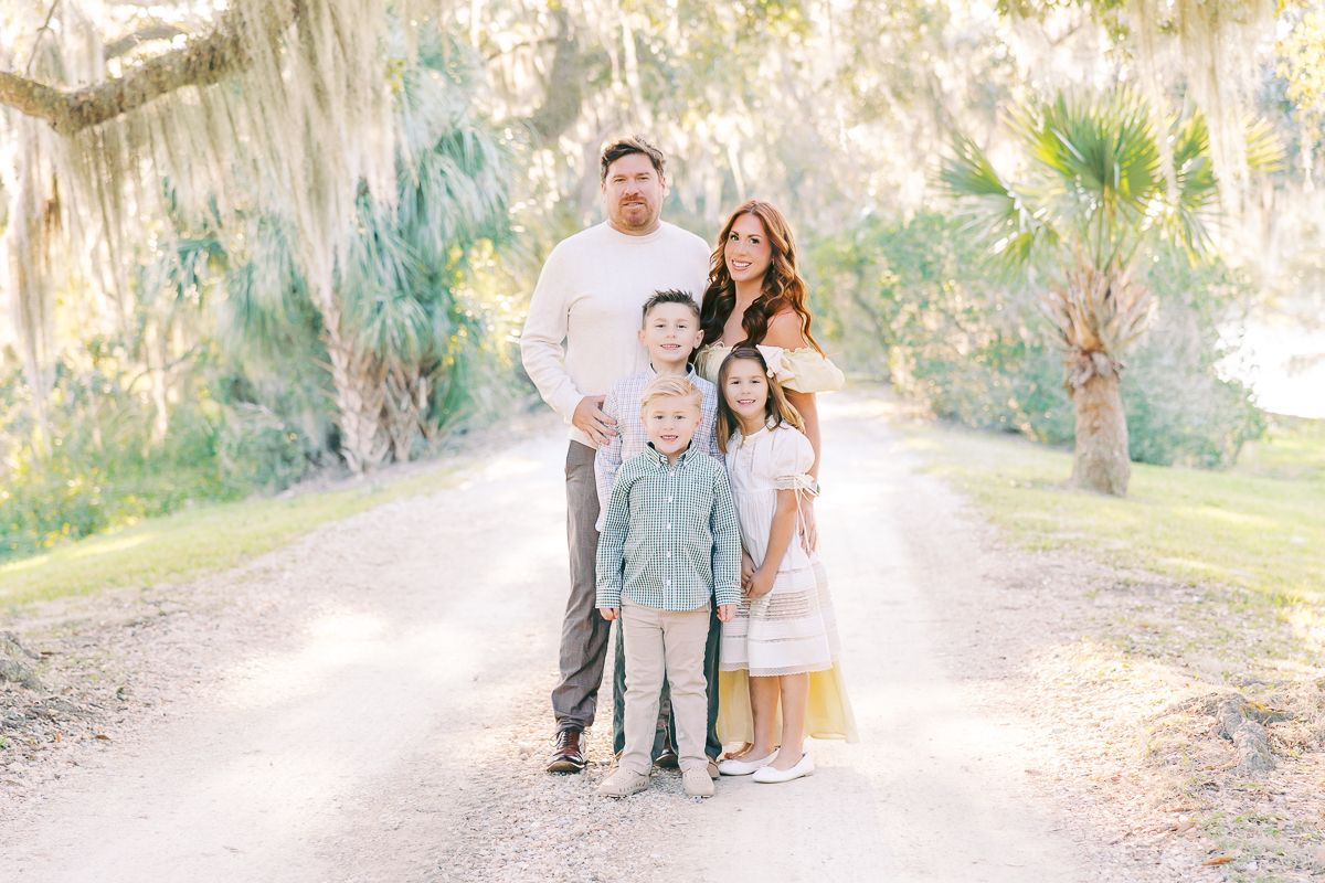 Family of five posing on a sunlit path surrounded by moss-draped trees in a bright outdoor portrait.