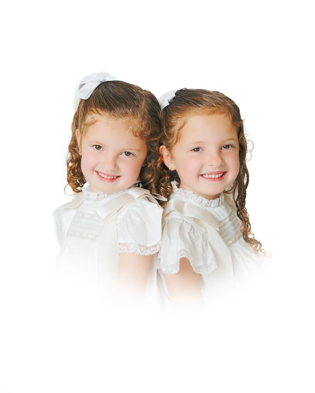 Two smiling young girls with curly hair wearing white dresses and hair bows pose back-to-back for a portrait photo.