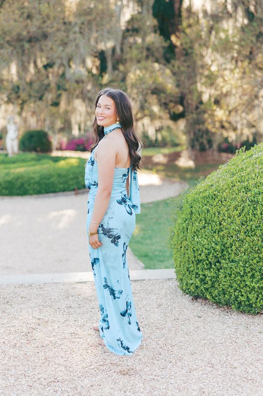 Woman in light blue floral halter maxi dress posing outdoors in a lush garden setting with trimmed hedges.