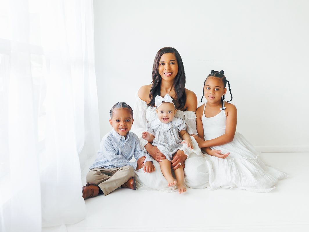 A woman sits with three young children in a bright white studio, all dressed in white and neutral tones.