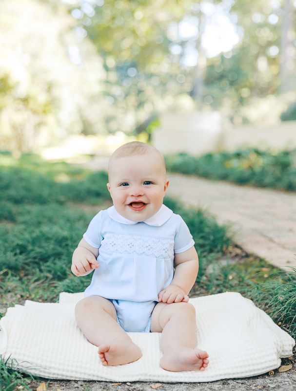Smiling baby in blue smocked romper sitting on white blanket outdoors on sunny day.