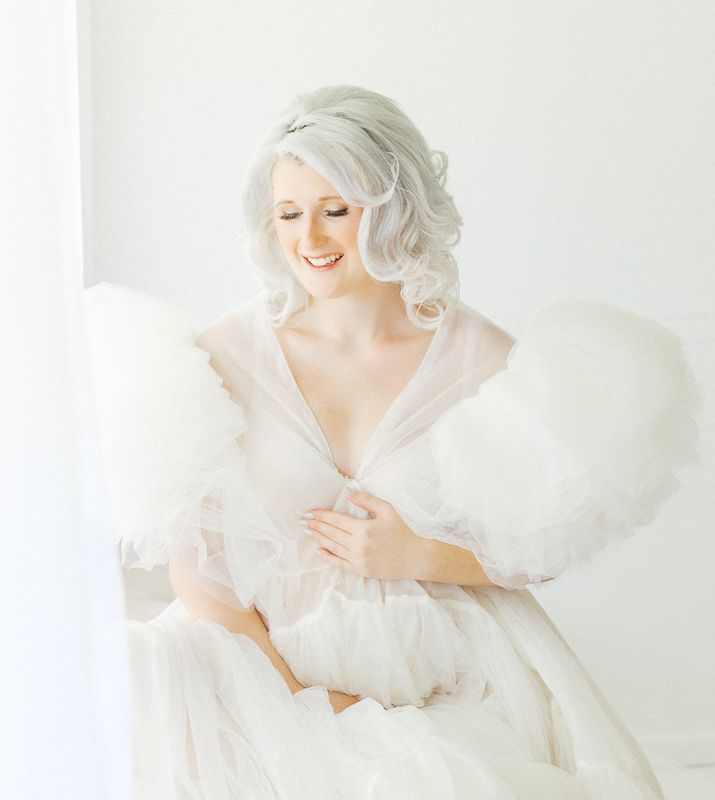 Elegant bride in a white gown with feather wrap smiling softly in a bright airy studio setting.