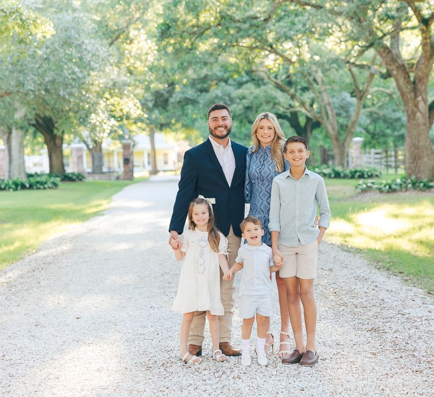 Family of five posing on a tree-lined gravel path, dressed in elegant neutral tones for a professional outdoor portrait.