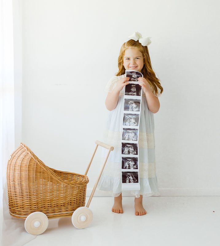 Red-haired girl in blue dress holds ultrasound strip beside wicker baby pram, announcing pregnancy.
