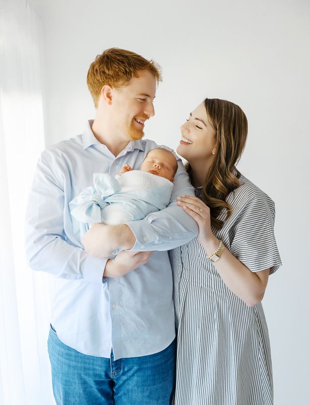 Smiling couple holding their newborn baby wrapped in blue, posing together in a bright white studio setting.