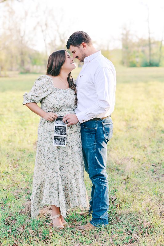 A couple shares a tender moment outdoors holding ultrasound photos, announcing their pregnancy in a sunlit field.