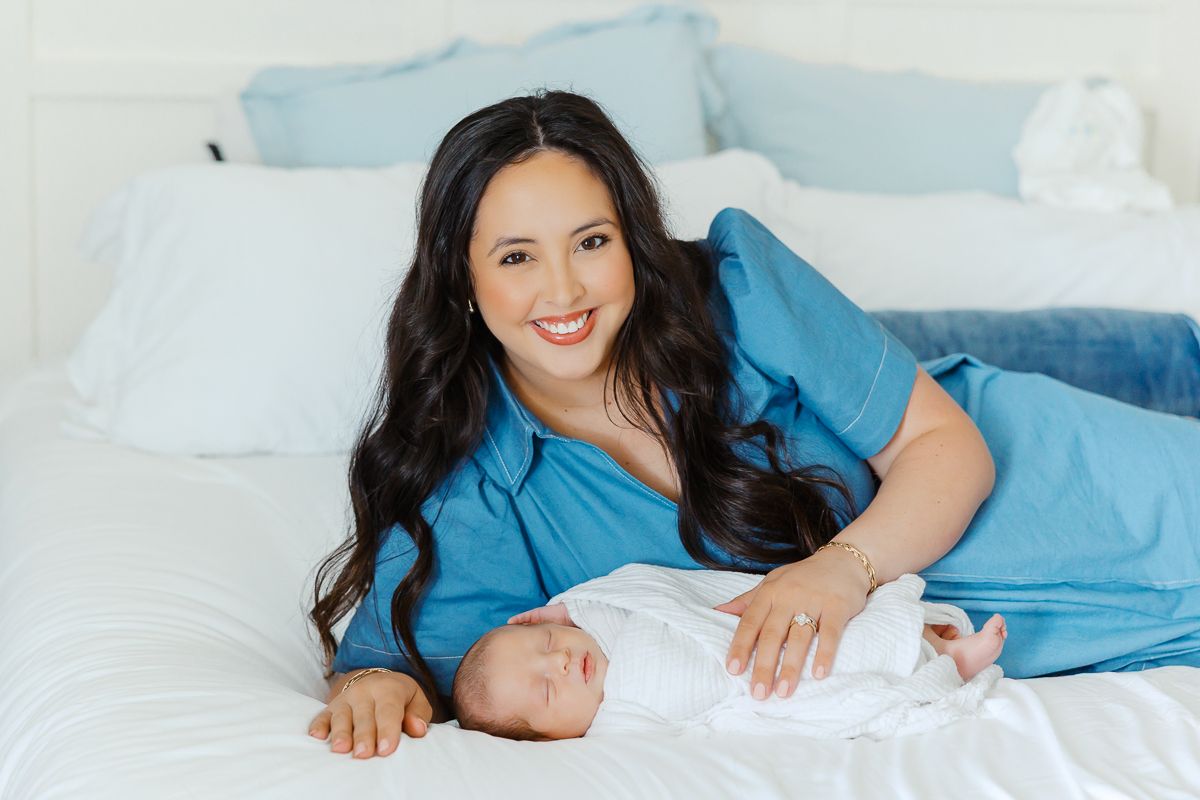A smiling woman in a blue dress lies on a white bed next to a sleeping newborn baby wrapped in white.