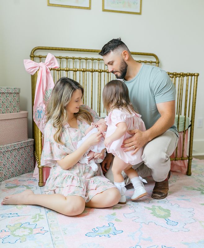 Family sitting on floor by gold crib in pink nursery, parents interacting with toddler girl in pink dress.