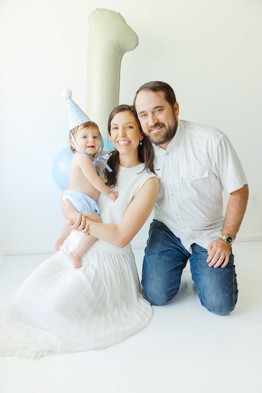 Family celebrating baby's first birthday with number one balloon and blue party hat in white studio.