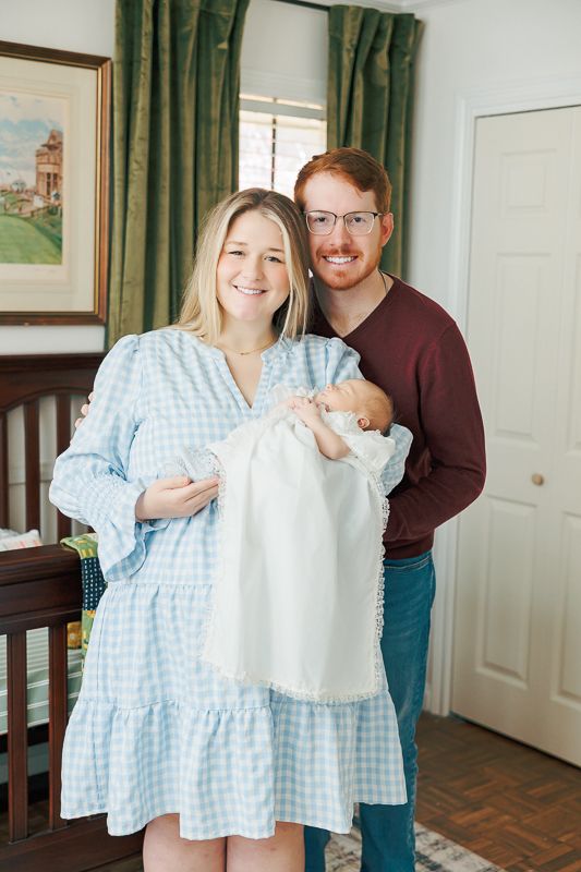 Happy couple smiling and holding their newborn baby in a white gown inside a cozy home nursery.