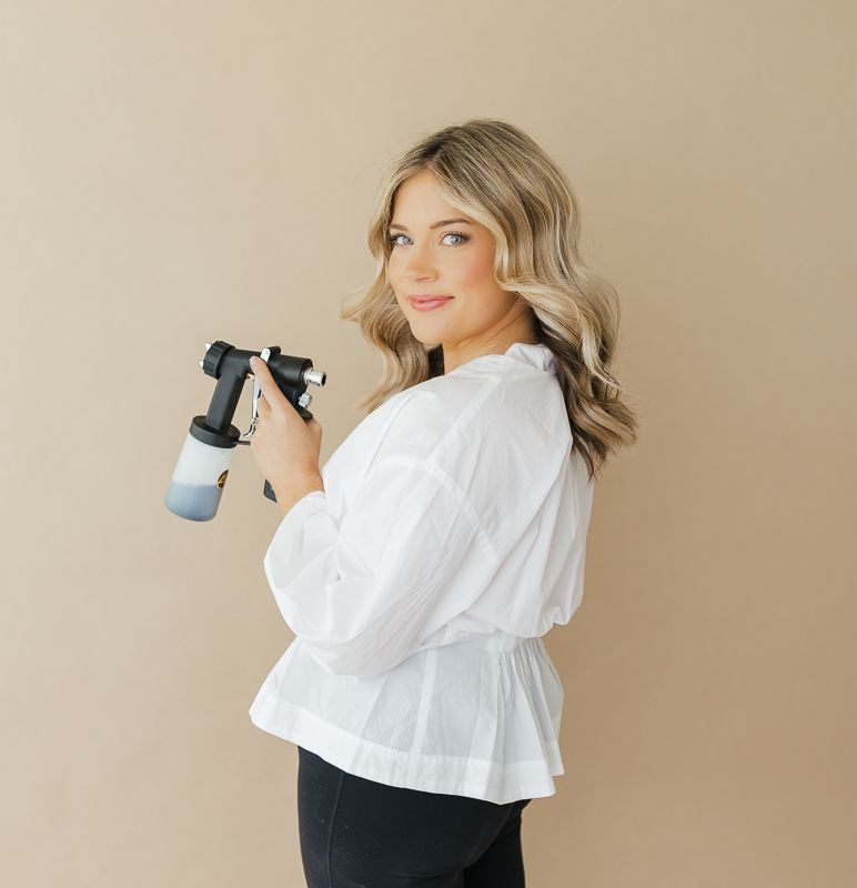 Woman in white blouse holding a massage gun, posing against a beige background.