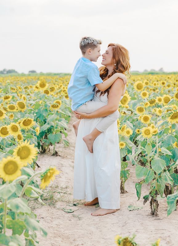 A mother holds her young son in a sunflower field, both smiling warmly in soft natural light.