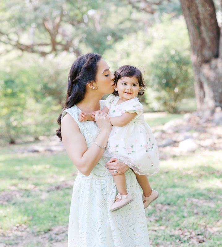 Mother kissing her smiling toddler daughter outdoors in matching white dresses among green trees.