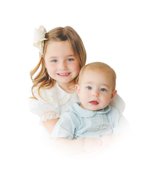 A smiling young girl with a bow in her hair hugs a baby boy in a light blue outfit against a white background.