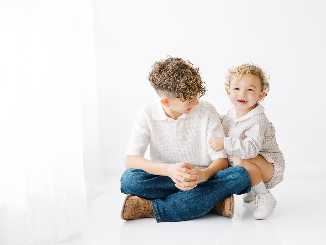 Two young brothers sit together smiling, older boy with curly hair crosses legs while toddler leans on him playfully.
