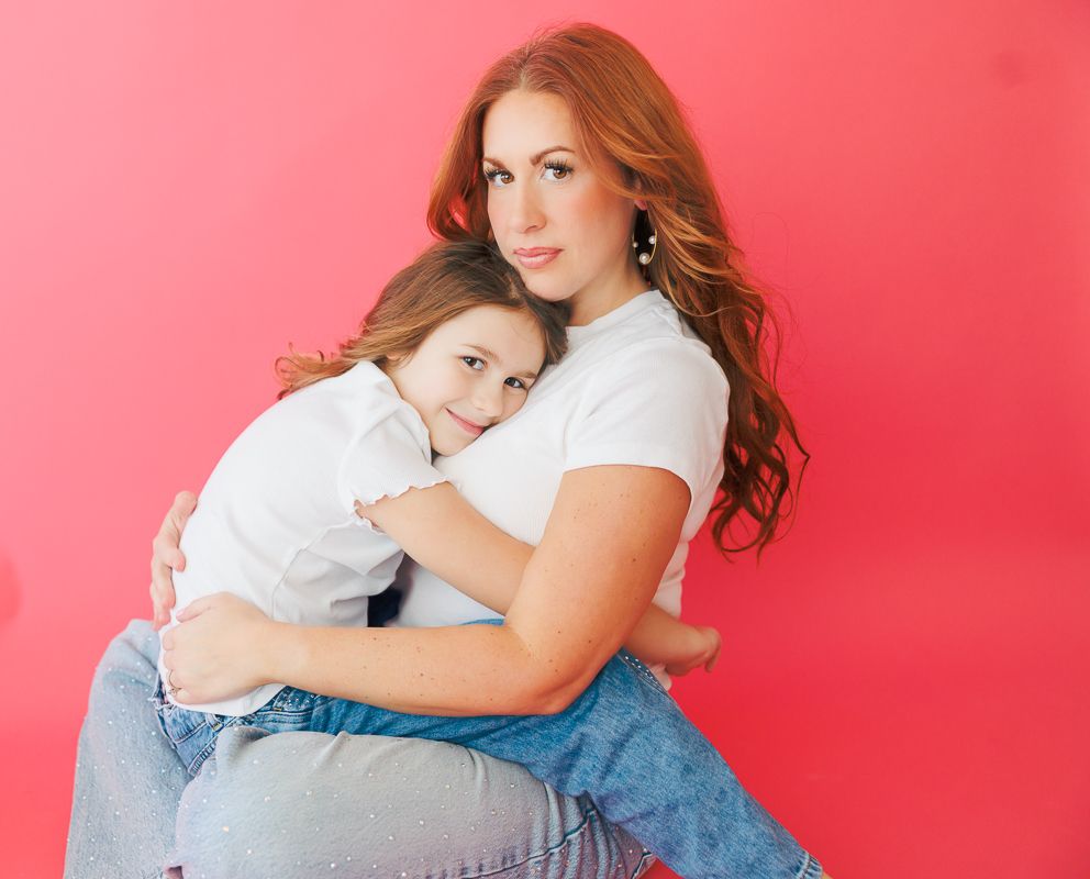 A woman with red hair hugs a young girl against a pink background, both wearing white tops and jeans.