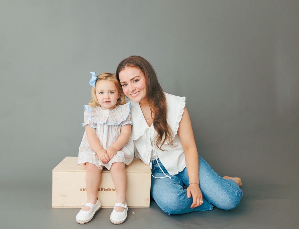 A young woman and toddler girl in matching light blue outfits pose together on a wooden box against a gray background.