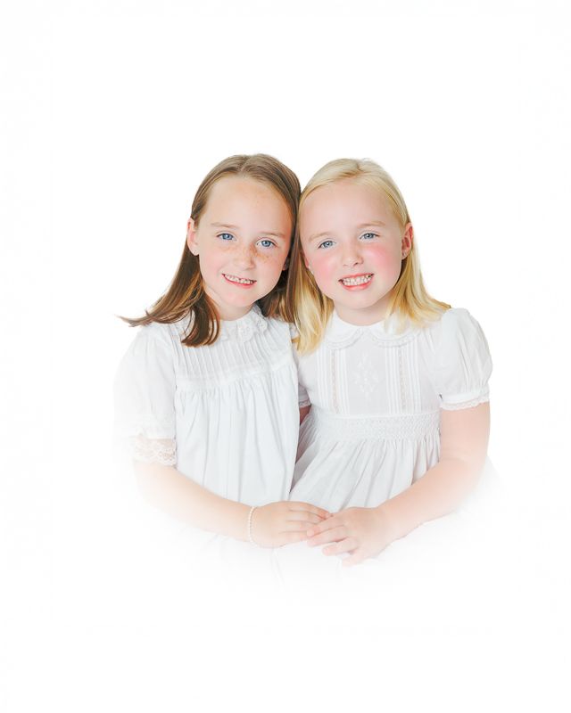 Two smiling girls in matching white dresses pose together for a portrait photo.