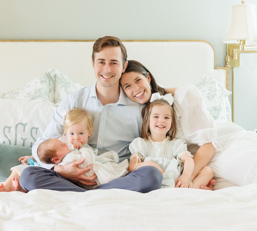 Happy family of five posing on a white bed, parents smiling with three young children including a newborn baby.