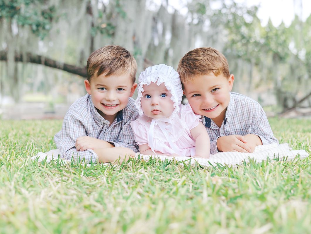 Two smiling boys and a baby girl in pink pose on grass under moss-draped trees in an outdoor family portrait.
