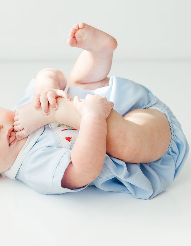 Adorable baby in blue outfit lying on back, playfully grabbing feet with chubby hands on white background.