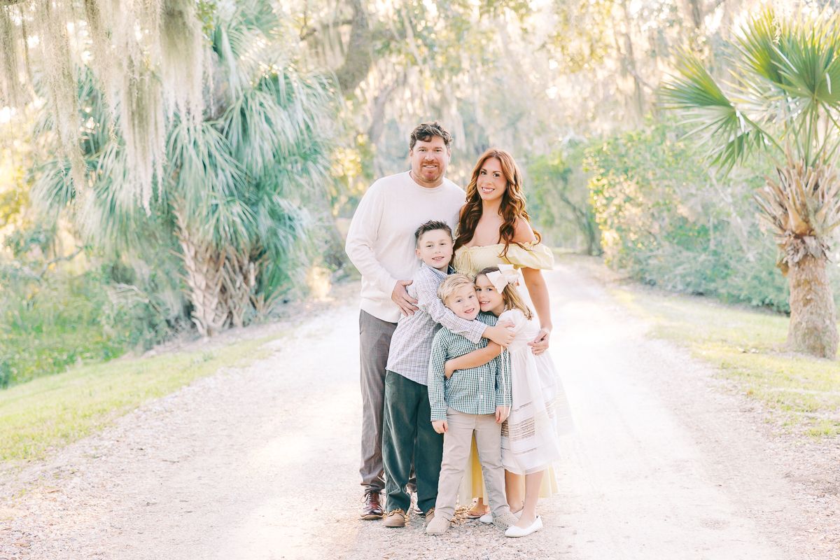 Family of four poses on a sunlit tree-lined path surrounded by lush greenery for an outdoor portrait session.