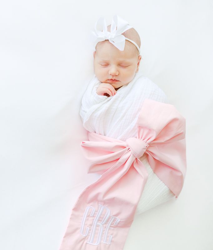 Sleeping newborn baby girl swaddled in white wrap with pink monogrammed bow sash and white bow headband.