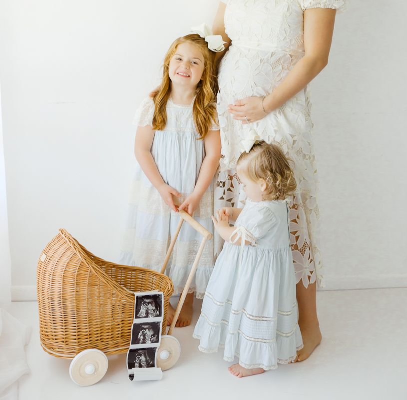 Pregnant mother with two young girls in white dresses posing with a wicker pram and ultrasound photos.
