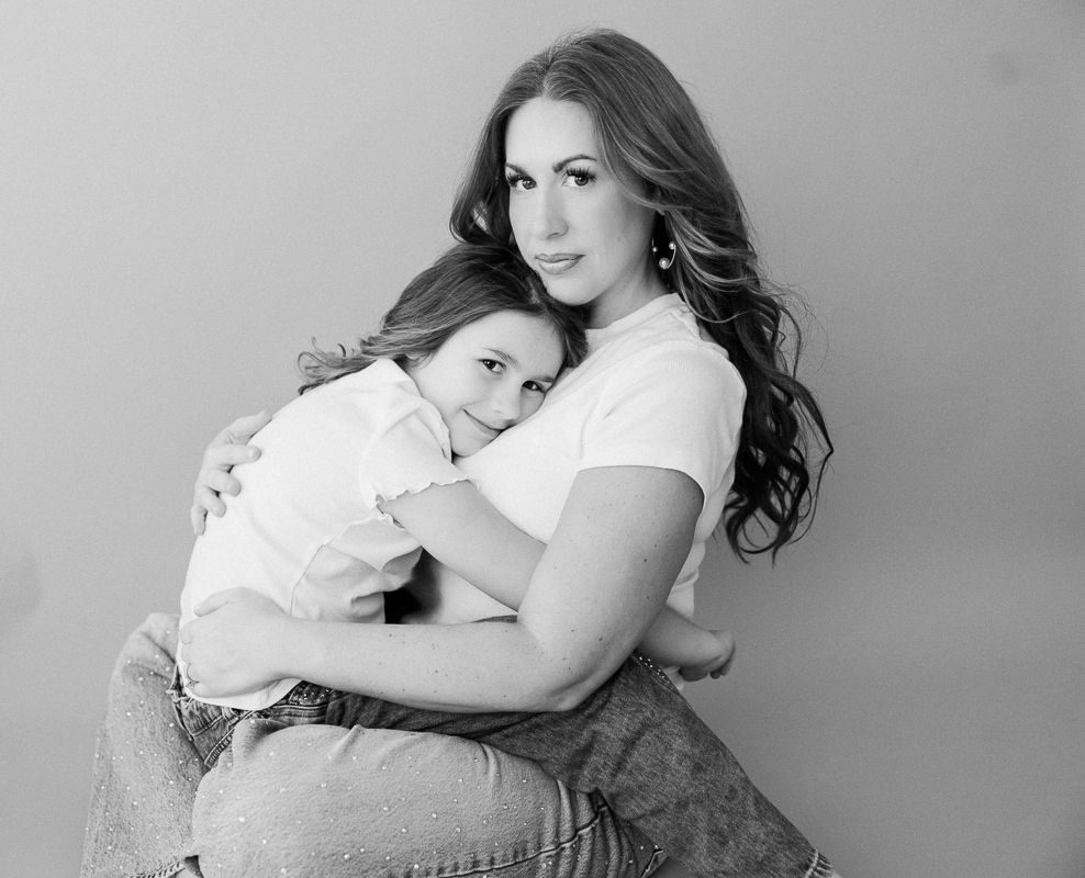 Black and white photo of a woman with long wavy hair hugging a young girl in a studio portrait.