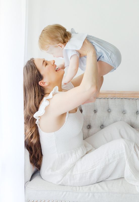 A smiling mother lifts her happy baby in the air while sitting on a cozy sofa in a bright white room.