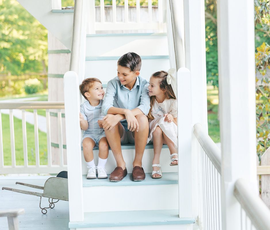 A man sits on white porch steps with two young children, smiling together in a warm, sunny outdoor setting.