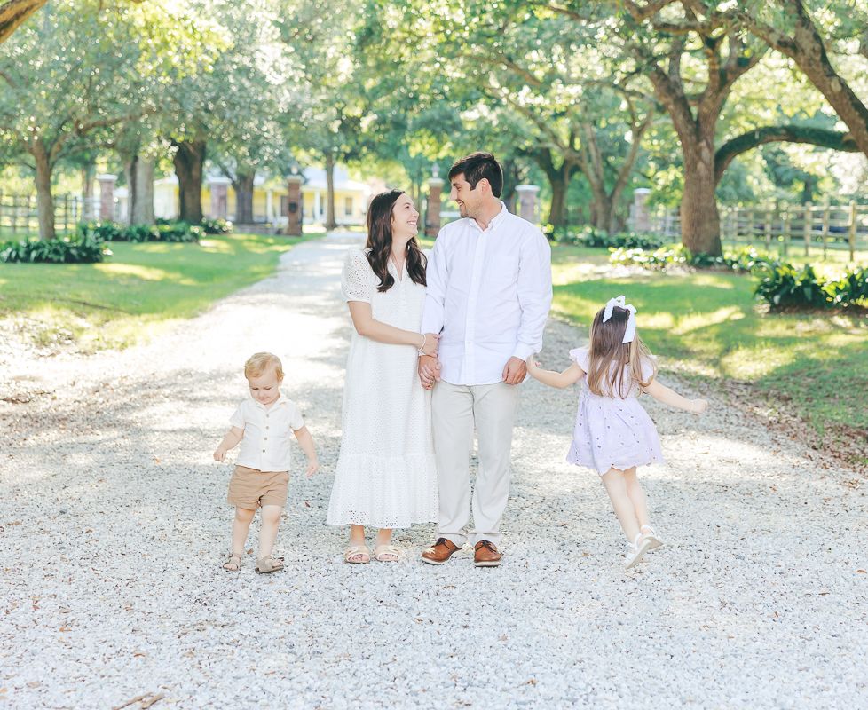 Family of four in white outfits walking on a tree-lined gravel path, parents holding hands with two young children.