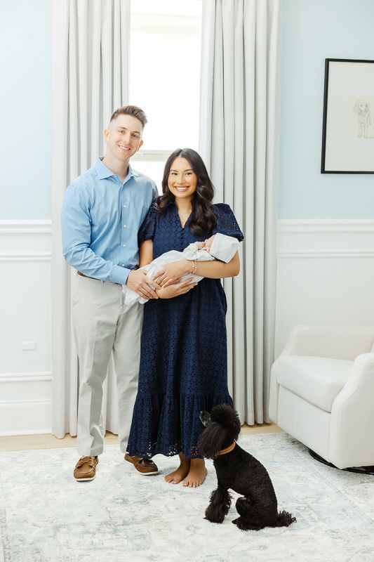 Happy couple holding newborn baby with black dog at their feet in elegant blue and white living room.