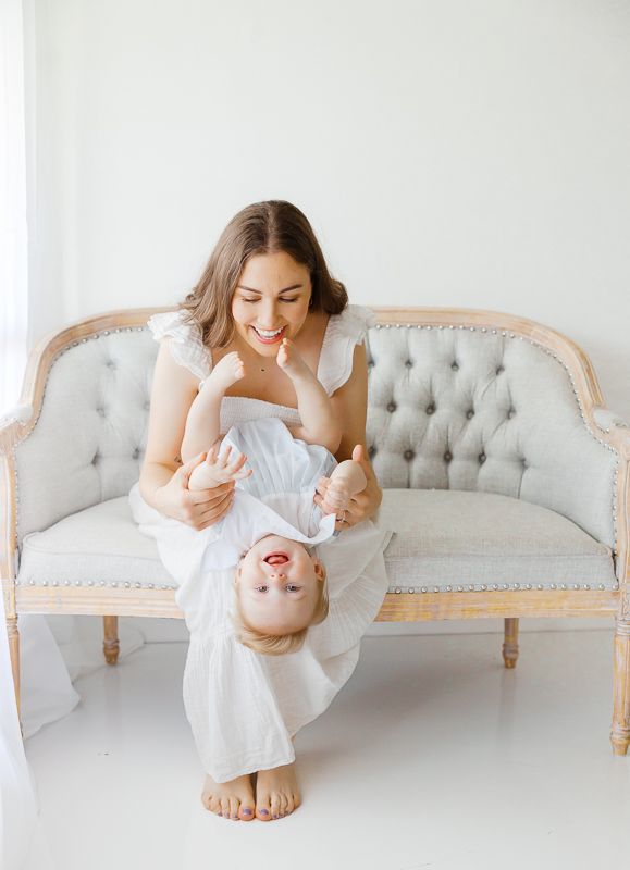 Smiling mother holding baby upside down on elegant tufted gray sofa in bright white room.