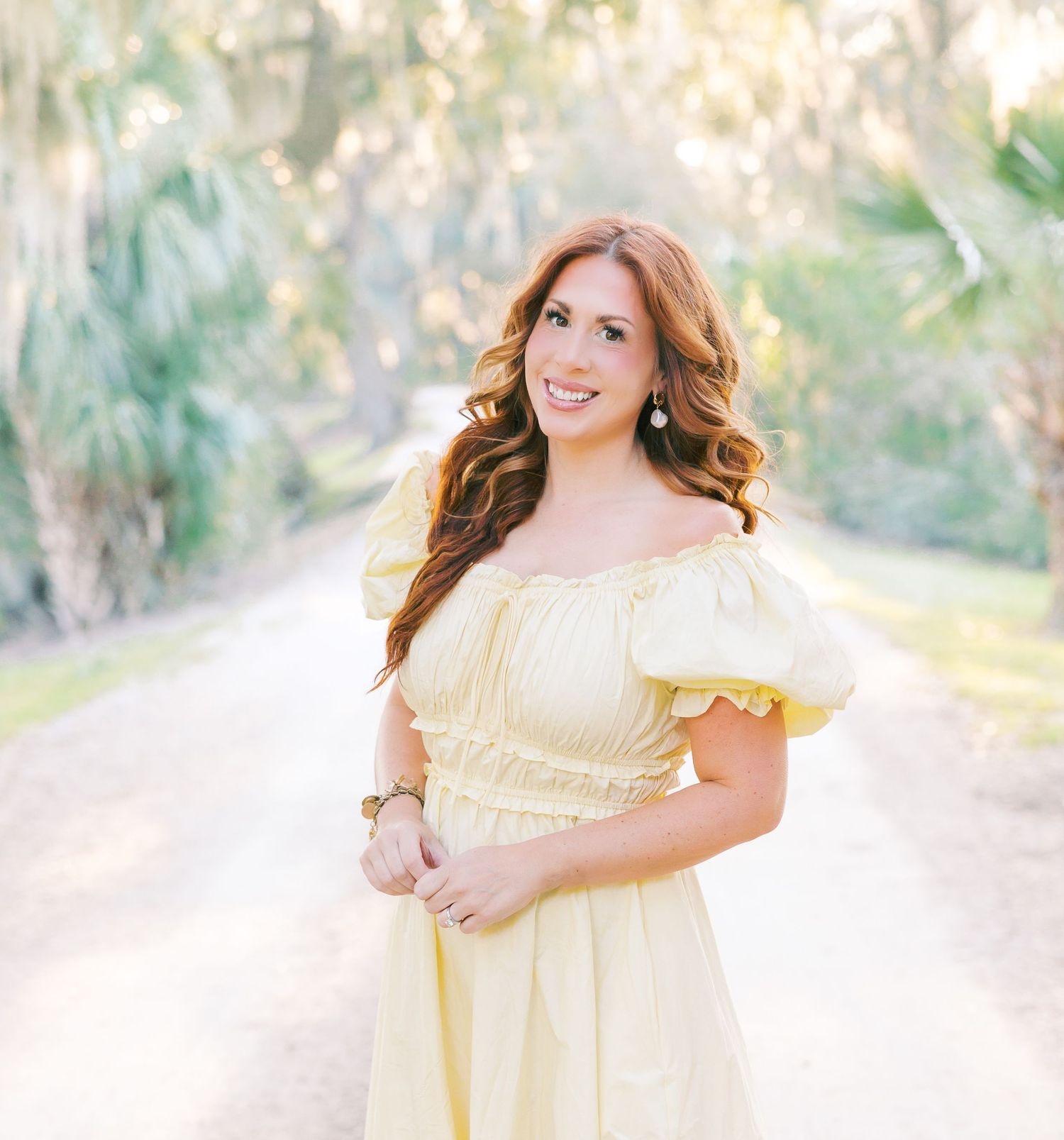 Smiling woman in yellow off-shoulder dress standing on tree-lined path in soft light.