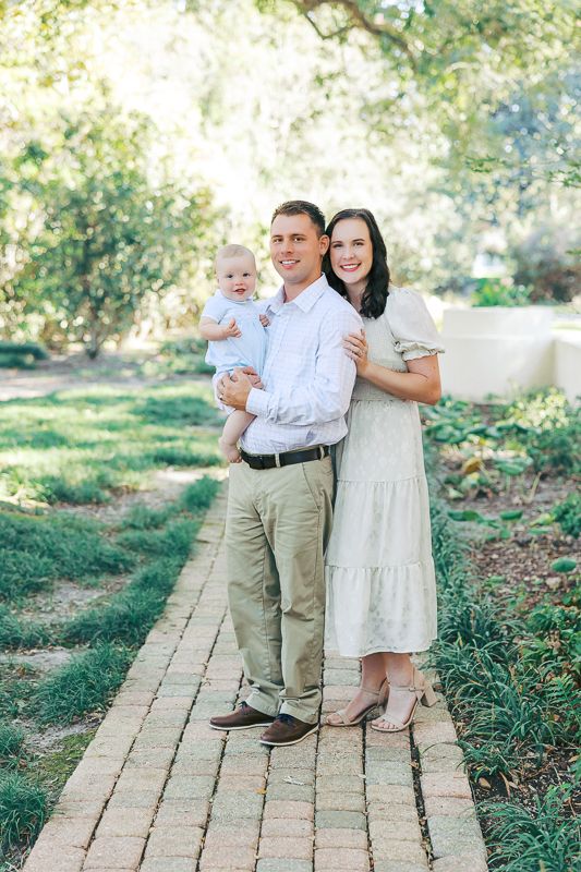 A young family poses on a brick path in a lush garden, parents smiling with their baby in a light blue outfit.