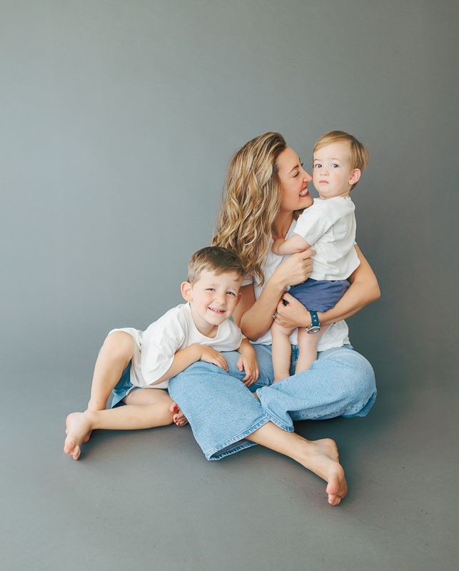 A mother sits on a gray studio floor in jeans, smiling and cuddling her two young red-haired boys in white shirts.
